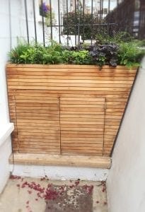 Rectangular wooden planter with horizontal slats, filled with various green plants and situated against a wall near outdoor storage units. Red flower petals are scattered on the ground nearby.