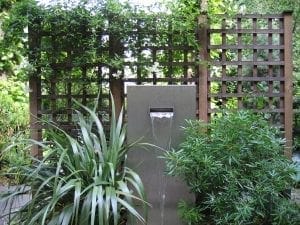 A modern rectangular water fountain stands in front of wooden lattice panels, surrounded by lush green plants, creating a serene garden setting ideal for relaxing before or after your health screening.