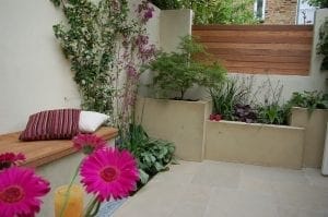 Small modern patio garden with beige walls, built-in wooden bench, a striped pillow, flowering plants, and greenery in raised beds; clever screening options add privacy—pink flowers and a drink brighten the foreground.