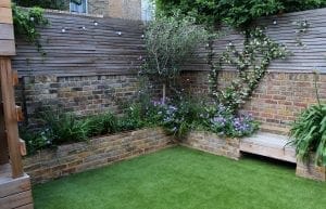 A small backyard garden with artificial grass, brick walls, wooden fencing, benches, and flower beds with purple and white blooms showcases creative screening options for added privacy.