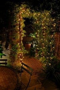 A garden at night with a vine-covered archway decorated with string lights, a round table and chairs in the foreground—an inviting spot to discuss top 10 screening options as you follow the gravel path leading further in.