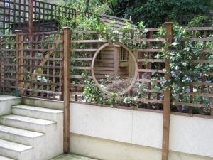 Wooden trellis fence with a circular opening, climbing plants, and a small wooden shed visible through the opening; stone steps are on the left—an inviting spot that could make any top 10 screening of garden privacy or creative screening options.
