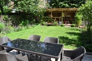 A backyard with a glass-top outdoor dining table and six chairs on a patio, a lawn, surrounding gardens, and a wooden pergola structure in the background—perfect for gathering to discuss top 10 screening options in comfort and style.