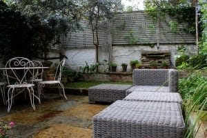 Outdoor patio with wicker seating, a metal table and chairs, potted plants, and a stone pathway—perfect for discussing top 10 screening options—enclosed by a wooden fence and greenery.