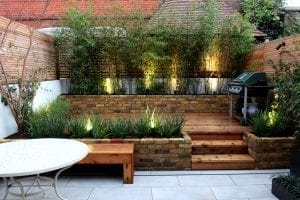 Small urban backyard with raised wooden deck, built-in brick planters, bamboo plants offering excellent screening options, spotlights, a barbecue grill, wooden bench, and round white table on tiled patio.