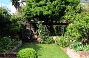 A neatly landscaped garden with a brick patio, green lawn, potted plants, flower beds, and a large leafy tree offering natural screening options for privacy and shade.