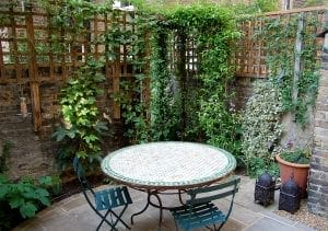 A small patio garden with a round mosaic table, two metal chairs, potted plants, climbing vines on trellises, and brick walls—an ideal spot to discuss top screening tests or explore various screening options in a relaxed setting.