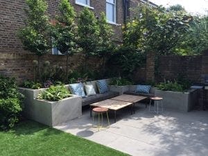 Modern patio with concrete bench seating, patterned cushions, a wooden coffee table, two small stools, lush greenery, and brick walls in a residential backyard offer inviting spaces to relax and discuss top screening tests and screening options.
