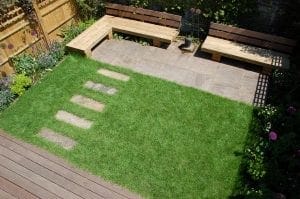 A small backyard showcases smart designs for small spaces, featuring a paved patio with wooden benches, a green lawn with stone stepping paths, and lush surrounding garden plants.