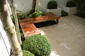 A modern L-shaped wooden bench is set against a white wall in a tiled garden patio, showcasing smart designs for small spaces and surrounded by trimmed shrubs and green plants.