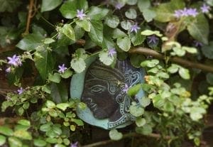 A round decorative plaque with a woman's profile is partially hidden among green leaves and small purple flowers, adding charm to small spaces with its smart design.