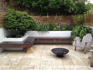 A modern patio with smart designs features a built-in wooden bench, potted plants, a fire pit, an Adirondack chair, and tiled flooring—perfect for small spaces and surrounded by greenery and a wooden slat fence.