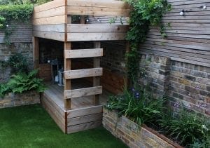 A wooden play structure with a ladder sits in a small backyard with artificial grass, brick walls, and surrounding plants—an example of smart designs perfect for small spaces.