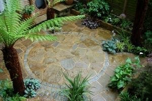A small garden patio with a circular stone-paved area, surrounded by various green plants and ferns—proof that smart designs can transform small spaces into tranquil retreats.