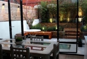 A modern dining area with a glass table and chairs overlooks a landscaped patio garden designed for small spaces, featuring wooden benches, brick planters, and ambient lighting.