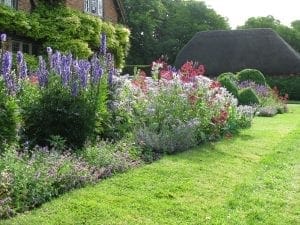 A well-maintained garden with neatly trimmed grass, colorful flowering plants, and bushes showcases beautiful planting combinations in front of a brick house and a thatched-roof building.