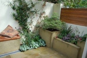 Smart designs make this small patio garden ideal for small spaces, with potted plants, a bench with a cushion, and light-colored walls partially covered by climbing plants.