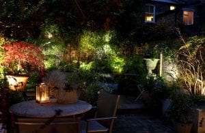 A dimly lit garden patio at night with a round table, chairs, potted plants, and a lantern—showcasing smart designs for small spaces—while illuminated greenery lines the stone wall.