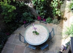 A round mosaic table with six chairs is set on a stone patio surrounded by leafy plants and a brick wall, showcasing smart designs perfect for small spaces in your garden.