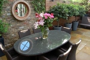A glass-top outdoor dining table with wicker chairs holds a vase of pink flowers; smart designs like potted plants and a round mirror maximize style in this patio area, making it perfect for small spaces.