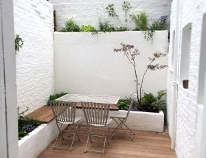 A small outdoor patio with a metal table and three chairs, surrounded by white brick walls and raised planters with various green plants—one of the top 10 smart designs for small spaces.