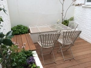 A small patio designed for small spaces, featuring a wooden deck, a white metal table with two chairs, and lush green plants against crisp white walls—perfect inspiration for smart designs in your home.
