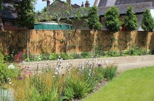 A neatly maintained garden with green lawn, flowering plants, and small trees, bordered by a brick wall and wooden fence, with buildings in the background.
