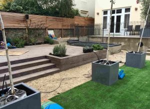 A modern backyard with artificial grass, large potted trees, a gravel path, wooden decking, and outdoor seating next to a house. Gardening supplies are scattered around.