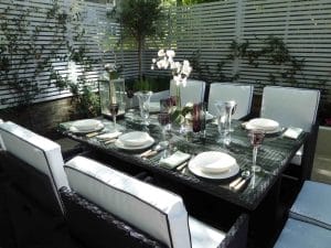A modern outdoor dining area with a glass-top table set for six, featuring white cushioned chairs, plates, glasses, and a flower centerpiece.