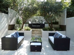 Modern outdoor patio with two black wicker sofas, a glass coffee table, and a dining area on a raised platform surrounded by trees and white slatted privacy screens.