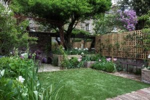A garden with a mix of green grass, stone paths, blooming flowers, leafy trees, and wooden fences surrounding the area.