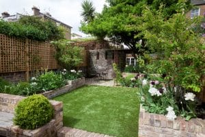 A small garden with a lawn, brick flowerbeds, blooming flowers, leafy trees, and a patio area with a table and chairs against a brick wall.
