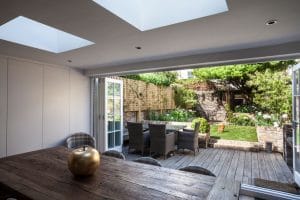View from a modern dining room with a wooden table and gold apple ornament, looking out through open glass doors onto a patio and garden with outdoor seating and greenery.