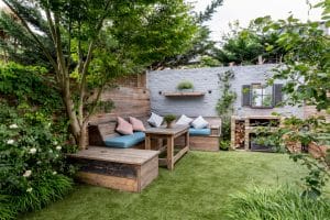 A cozy backyard patio with wooden benches and tables, blue cushions, green plants, and a gray brick wall with shelves, firewood, and a mirror.