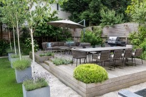 A modern outdoor patio with a wooden deck, dining table and chairs, lounge area, umbrella, potted plants, and a barbecue grill, surrounded by greenery and fencing.