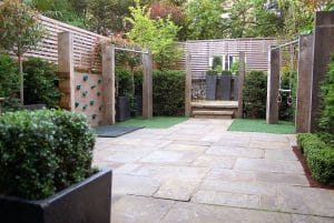 A paved backyard with a mini climbing wall, gym rings, artificial turf, and wooden fencing, surrounded by green shrubs and small trees.
