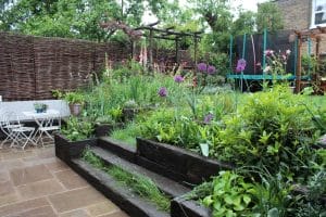 A tiered garden with green plants and purple flowers, stone steps, a white outdoor table and chairs, a trampoline, and a woven fence in the background.