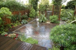 A small, modern garden with wet stone paving, wooden decking, lush green plants, and trellised wooden fences. A stainless steel water feature is visible in the corner.