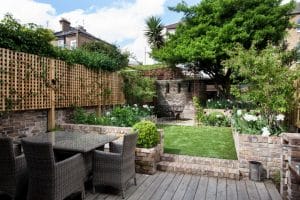 A tidy backyard with a wooden deck, wicker dining set, brick planters, green lawn, trees, and a wooden fence against a backdrop of neighboring buildings.