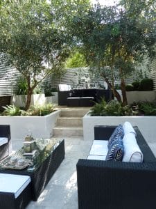 Modern outdoor patio with black wicker furniture, white cushions, blue accent pillows, glass coffee table, greenery, and trees surrounding a raised seating area.