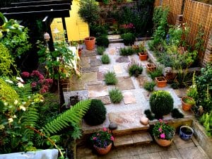 A neatly arranged garden with stone paths, potted plants, trimmed bushes, flower beds, and a yellow shed in the background.