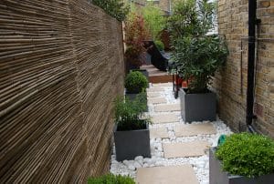 A narrow outdoor walkway with square stone pavers, white decorative rocks, potted green plants, a bamboo fence on one side, and a brick wall on the other.