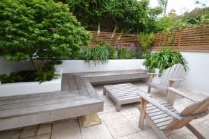 A small garden patio with wooden benches, two chairs, low tables, potted plants, and wooden slat fencing on white stone tile flooring.