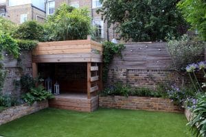 A small backyard with artificial grass, a raised wooden deck structure, brick walls, and surrounding greenery and flowers.