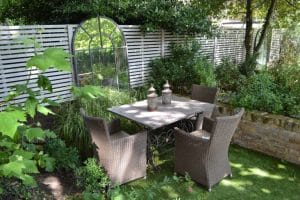 A garden patio with a square dining table, four wicker chairs, and three decorative lanterns, surrounded by lush green plants and a white slatted fence.