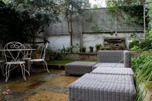 A small garden patio with wicker seating, a metal table and chairs, potted plants, and a wooden fence in the background.