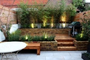 A small backyard garden with brick planters, wooden steps, bamboo plants, a green barbecue grill, and a round white table on a tiled patio.