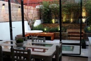 Modern dining area with glass doors opens to a landscaped patio featuring wooden benches, a round table, greenery, and up-lit garden beds against a wooden privacy fence.