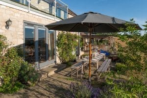 Outdoor dining area with a long wooden table and benches under a large umbrella on a stone patio beside glass doors, surrounded by lush garden plants—a true summer oasis perfect for relaxed gatherings.