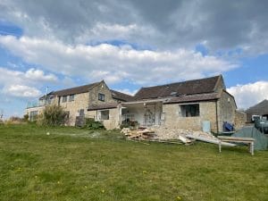 A stone house with an attached barn under renovation, debris and construction materials scattered in the grassy yard, hints at its potential as a summer oasis beneath a cloudy sky.